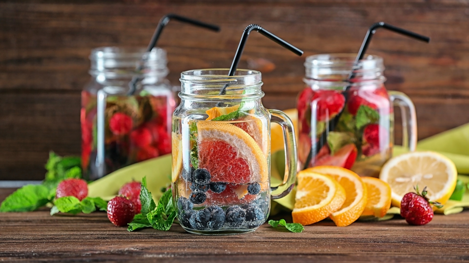 Mason jars of infused water with fruits on wooden table Drei Gläser mit fruchtinfusiertem Wasser, gefüllt mit Erdbeeren, Blaubeeren, Orangen- und Zitronenscheiben sowie Minzblättern.