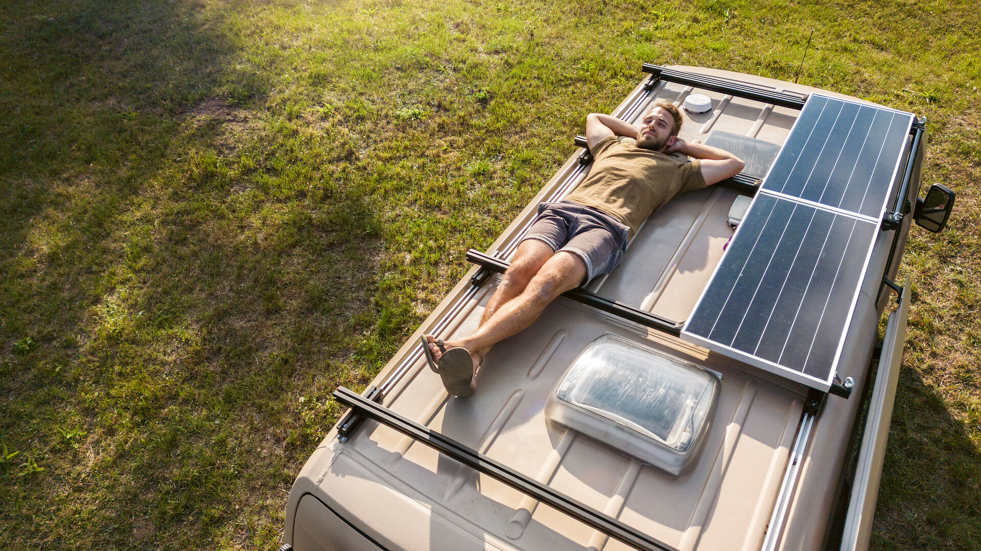 Mann liegt auf Wohnmobildach mit Solarpaneelen, geparkt auf grasbewachsener Fläche