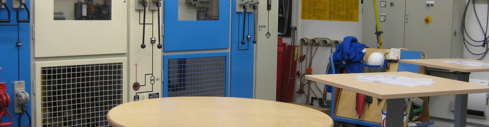 Raum mit Industrieausrüstung und Steuerpulten, Werkzeuge und Sicherheitsausrüstung im Vordergrund.