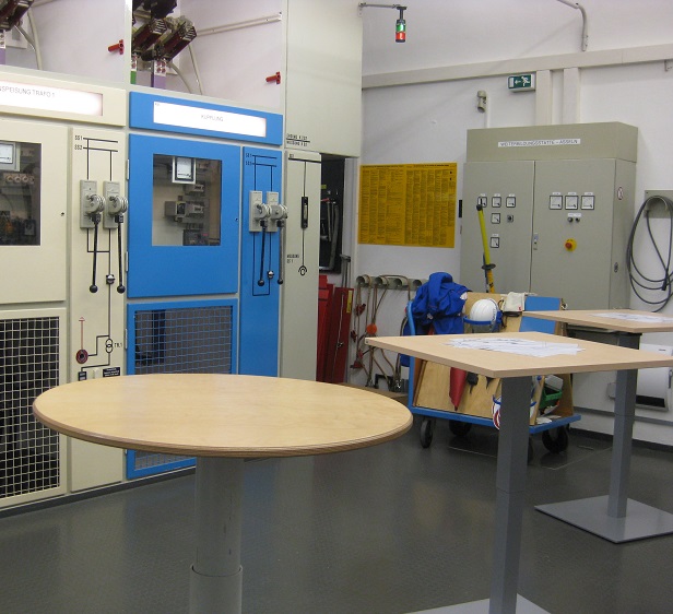 Raum mit Industrieausrüstung und Steuerpulten, Werkzeuge und Sicherheitsausrüstung im Vordergrund.
