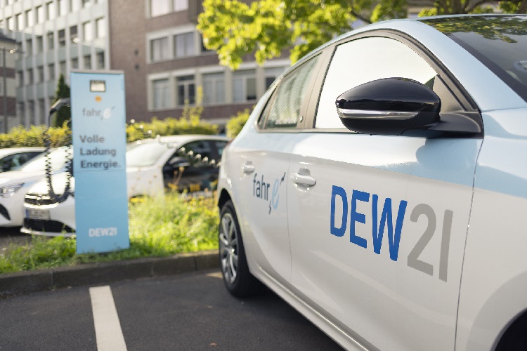 Nahaufnahme eines weißen Fahrzeugs mit 'DEW21' auf der Seite, weiteres Fahrzeug und Grün im Hintergrund
