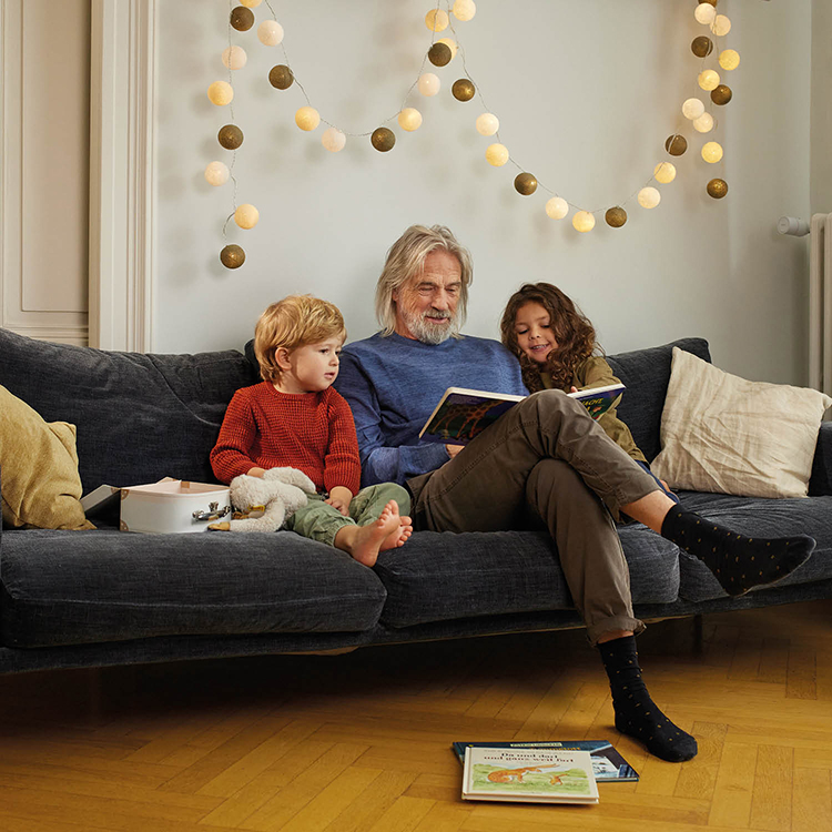 Drei Personen auf dunkelblauem Sofa, mittlere Person hält ein Buch, zwei schauen zu, Lichterkette an der Wand, Bücher und weiße Box auf dem Sofa, zwei Bücher auf dem Boden.