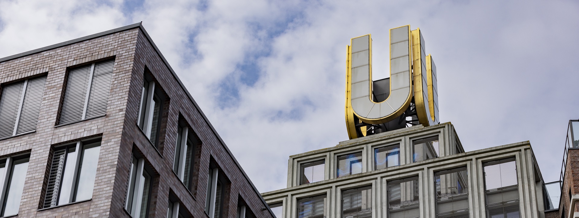 Modernes Gebäude mit großem, goldenem 'U' auf dem Dach, mehrere Etagen mit großen Fenstern, aus Ziegel und Glas, teilweise bewölkter Himmel.