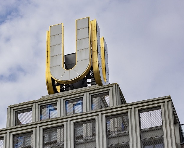Modernes Gebäude mit großem, goldenem 'U' auf dem Dach, mehrere Etagen mit großen Fenstern, aus Ziegel und Glas, teilweise bewölkter Himmel.