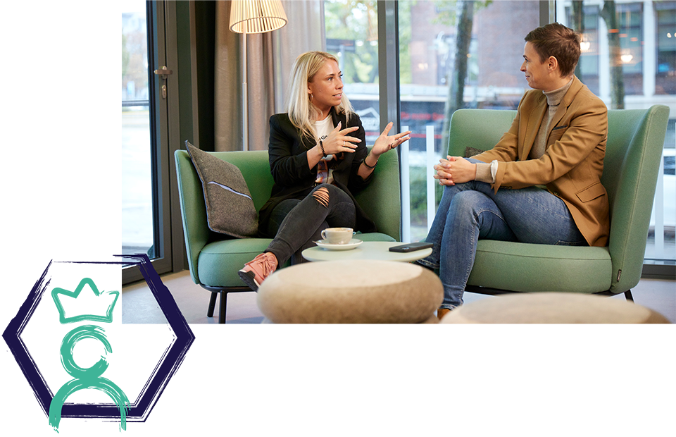 Zwei Personen auf grünen Sofas in modernem, gut beleuchtetem Raum mit großen Fenstern, Gespräch, Kaffeetasse und Untertasse auf dem Tisch, Symbol einer Person mit Krone in der unteren linken Ecke.