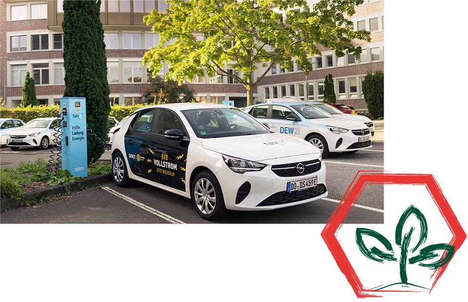 Parkplatz mit drei weißen Autos, Vordergrundauto mit Logo und Text 'WALLSTION' und 'DEW21', blaues Schild 'E-Mobility Ladepunkt', rotes Sechseck mit grünen Blättern unten rechts.