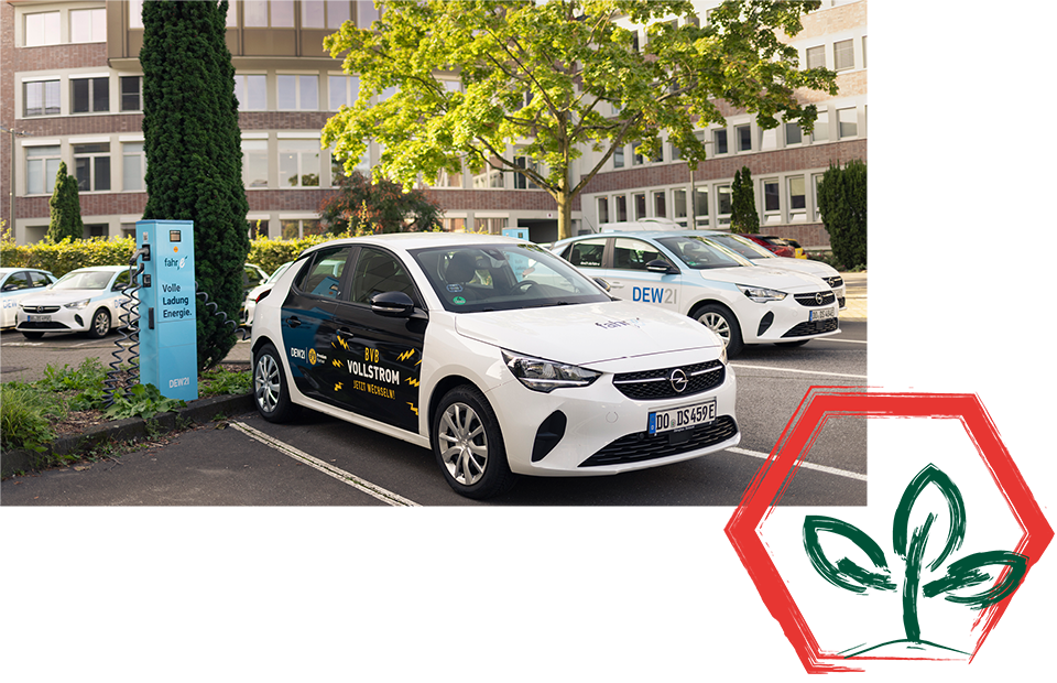 Parkplatz mit drei weißen Autos, Vordergrundauto mit Logo und Text 'WALLSTION' und 'DEW21', blaues Schild 'E-Mobility Ladepunkt', rotes Sechseck mit grünen Blättern unten rechts.