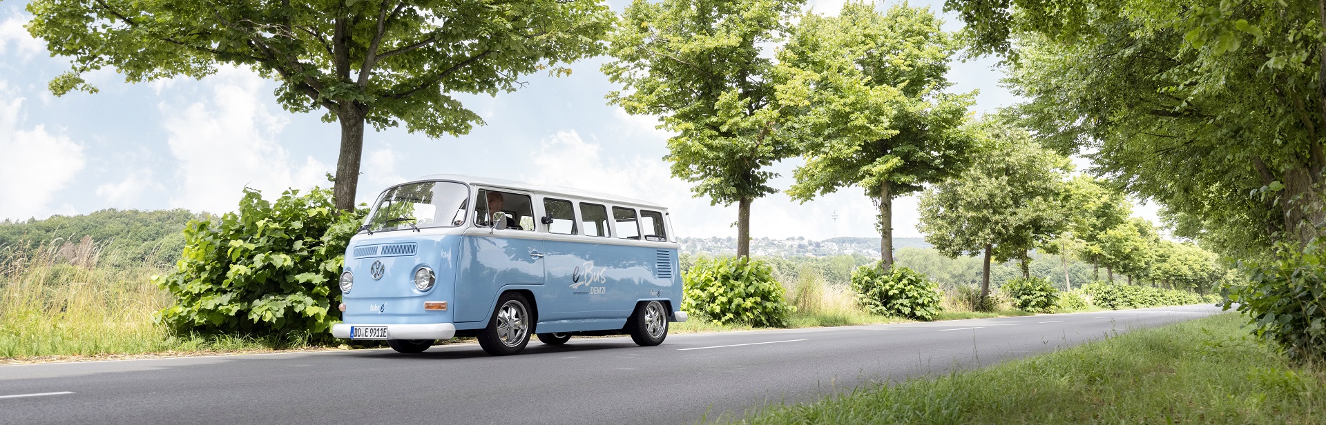 Hellblauer DEW21 E VW-Bus auf einer Straße, umgeben von Bäumen und Grün.