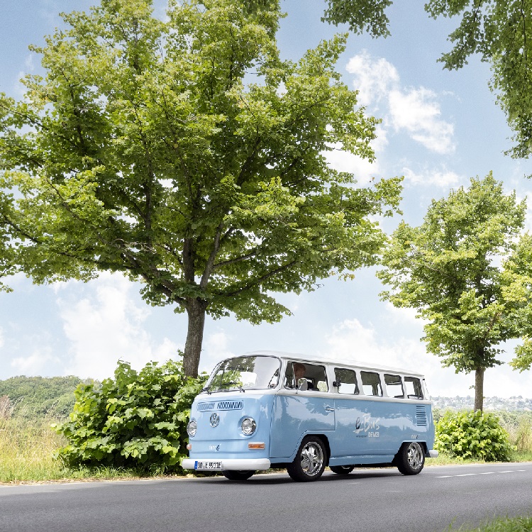 Hellblauer DEW21 E VW-Bus auf einer Straße, umgeben von Bäumen und Grün.