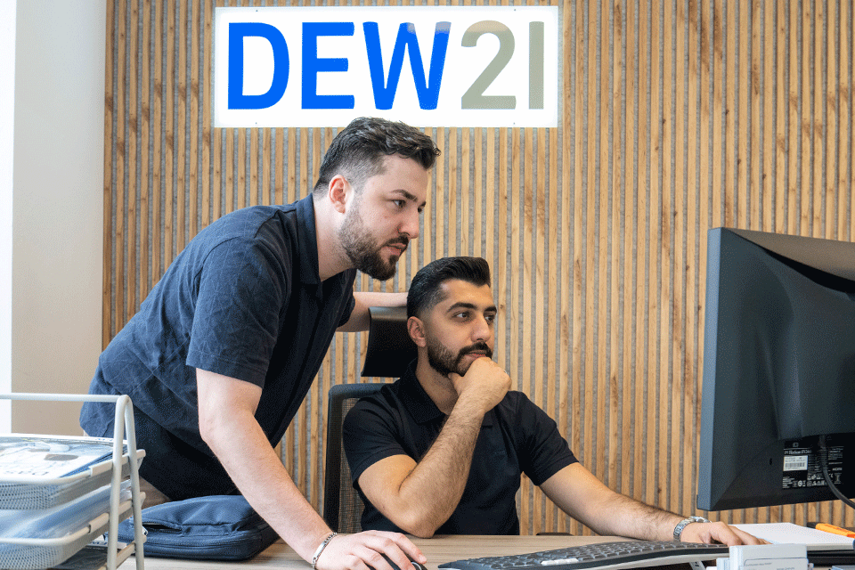 zwei männer sitzten vor einem Computerbildschirm im Hintergrund ist das dew21 logo an der Wand