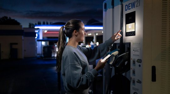 Eine Frau an einer beleuchteten Ladestation für Elektrofahrzeuge des Unternehmens "DEW21" zeigt auf den Bildschirm der Station. Es ist dunkel, und im Hintergrund ist eine Tankstelle zu sehen.