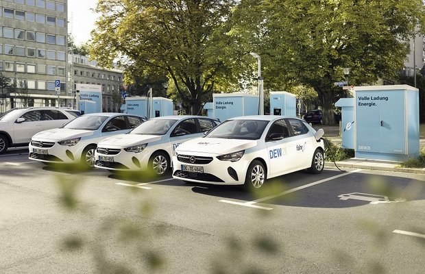 Drei weiße Elektroautos an blauen Ladestationen, Aufschrift 'DEW' auf Autos, 'Volle Ladung Energie' auf Ladestation.