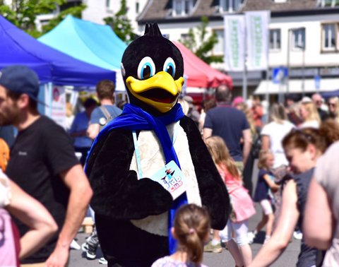 DEW21 Maskottchen Pido bei einer Veranstaltung, Menschen und Zelte im Hintergrund.