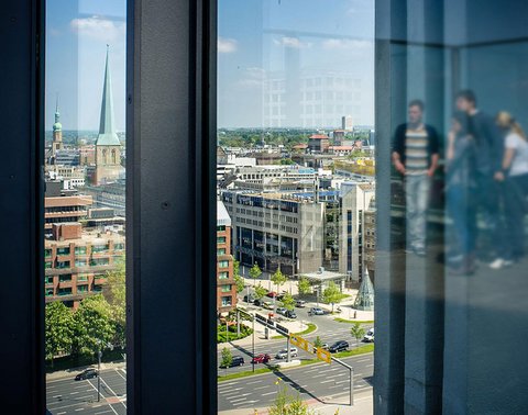 Stadtansicht durch ein großes Fenster mit der Spiegelung einer Gruppe von Menschen. Gebäude und Grünflächen im Hintergrund.