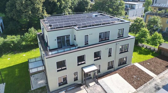Modernes Gebäude mit Flachdach und Solarpanels, umgeben von Grünflächen und Bäumen.