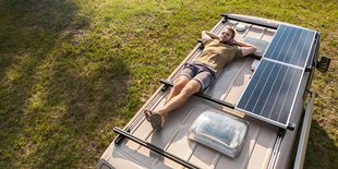 Person liegt auf Fahrzeugdach neben Solarpaneelen, geparkt auf Grasfläche.