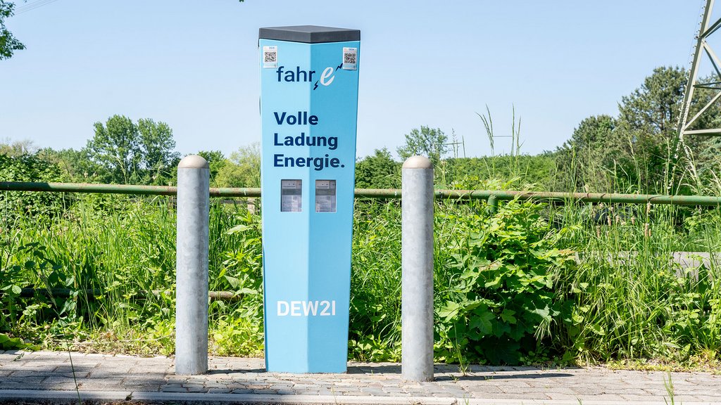 Blaue E-Ladesäule von DEW21 mit Werbeslogan, umgeben von Pollern und Vegetation