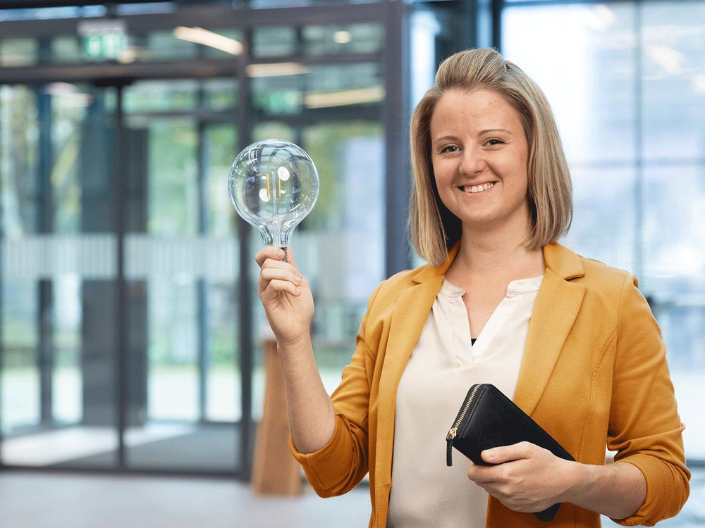 Person hält eine große Glühbirne und eine schwarze Geldbörse, trägt einen gelben Blazer.