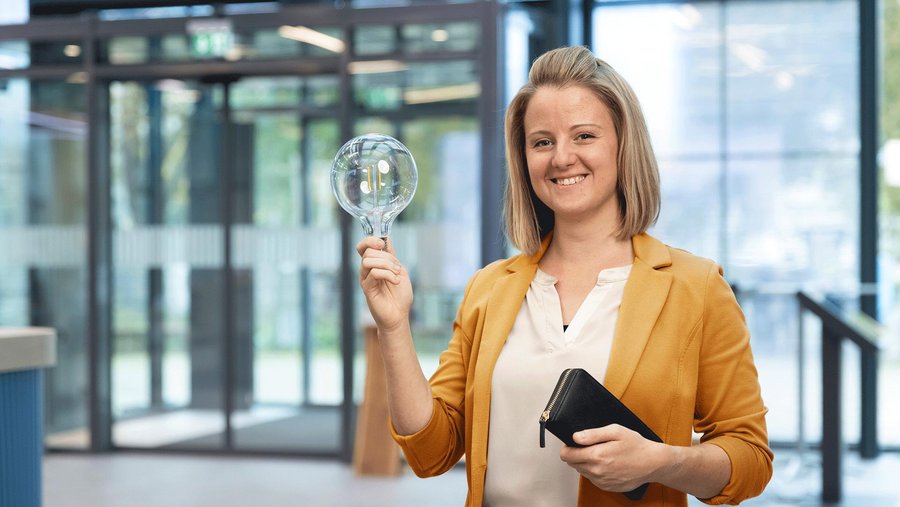 Person hält eine große Glühbirne und eine schwarze Geldbörse, trägt einen gelben Blazer.
