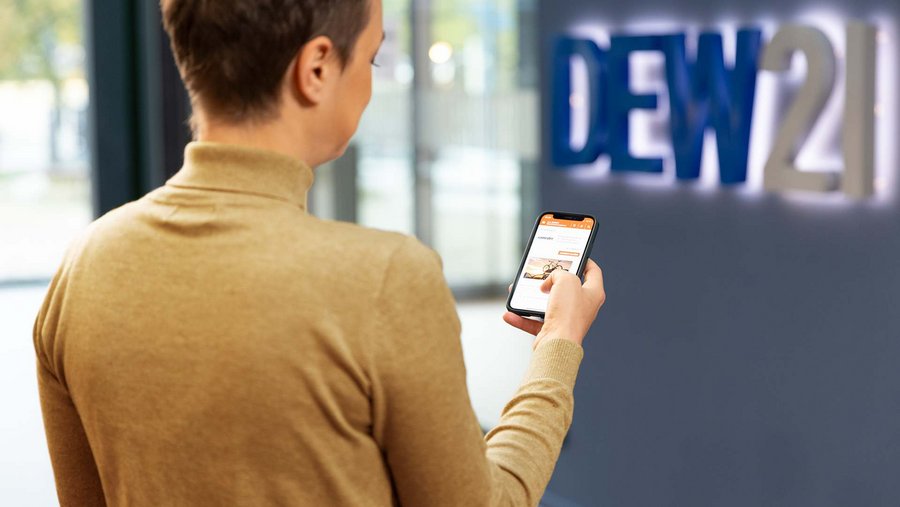 Person hält ein Smartphone, Hintergrund zeigt 'DEW21'