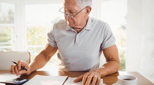 Ältere Person am Tisch mit Papieren, Stift und Taschenrechner, im Hintergrund helle Fenster.