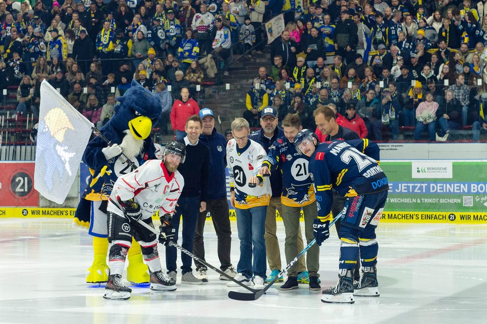 Zwei Eishockeyspielende beim Face-off auf dem Eis, umgeben von mehreren Personen und dem Adler-Maskottchen.