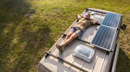 Mann liegt auf Wohnmobildach mit Solarpaneelen, geparkt auf grasbewachsener Fläche