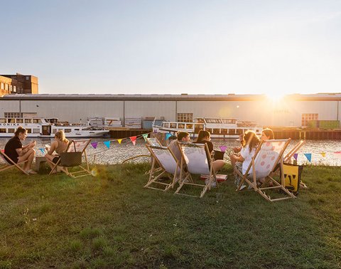 Menschen auf Liegestühlen an einem Gewässer bei Sonnenuntergang. Gebäude und Boote im Hintergrund.