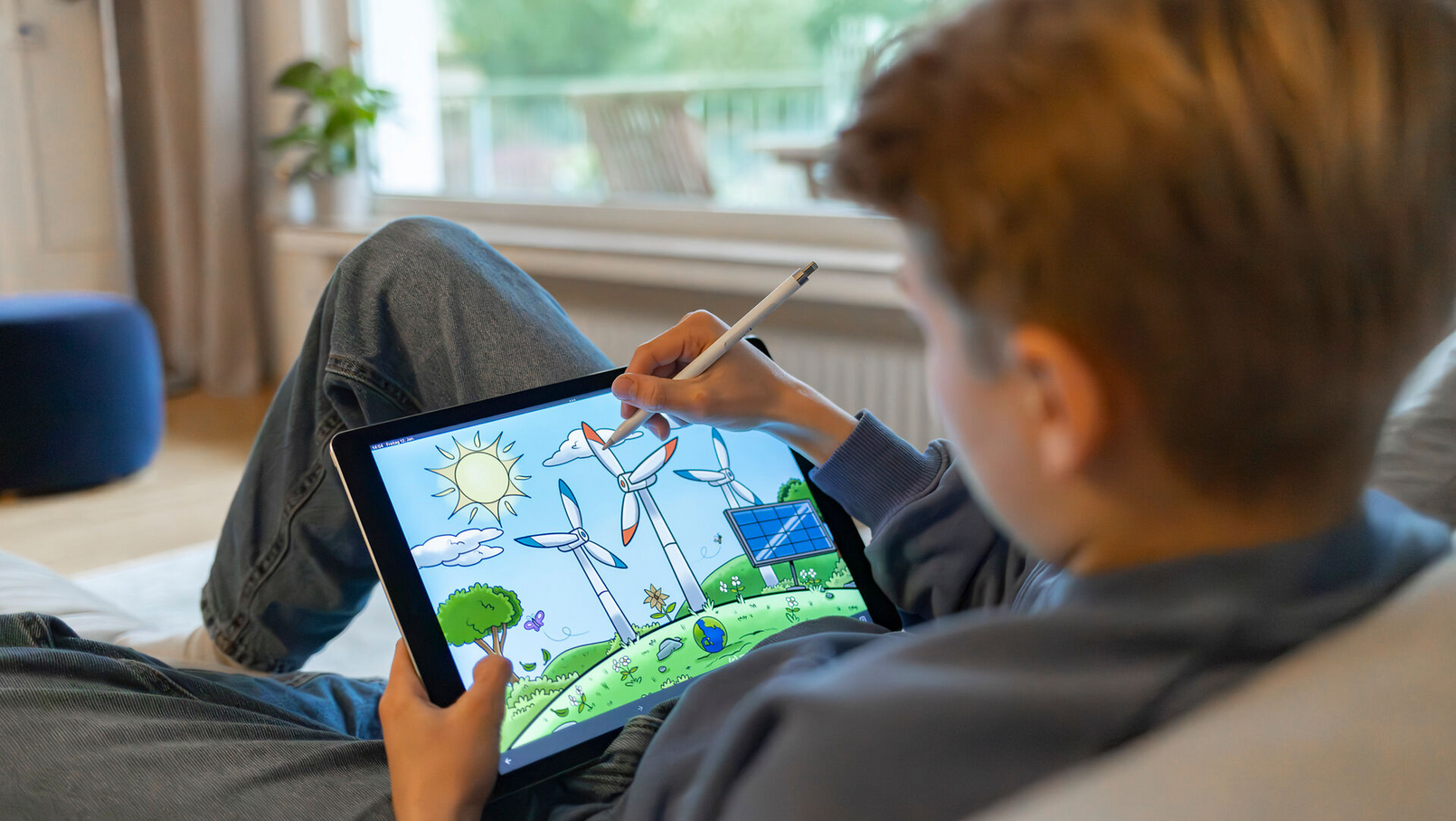 Kind zeichnet mit Stift auf Tablet eine bunte Landschaft mit Windrädern und Sonne