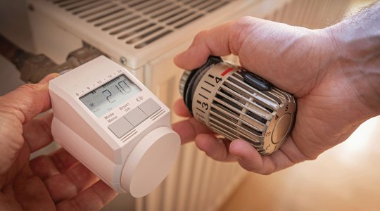 Zwei Hände arbeiten an einem Heizkörper. Die linke Hand hält ein digitales Thermostat, die rechte einen herkömmlichen Regler.
