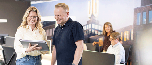 Mann und frau gucken auf tablet in einem Büro mit Menschen an Rechnern im Hintergrund