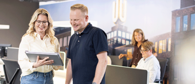 Mann und frau gucken auf tablet in einem Büro mit Menschen an Rechnern im Hintergrund