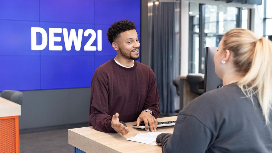 Zwei Personen interagieren an einem Schalter, im Hintergrund ein Bildschirm mit "DEW21".